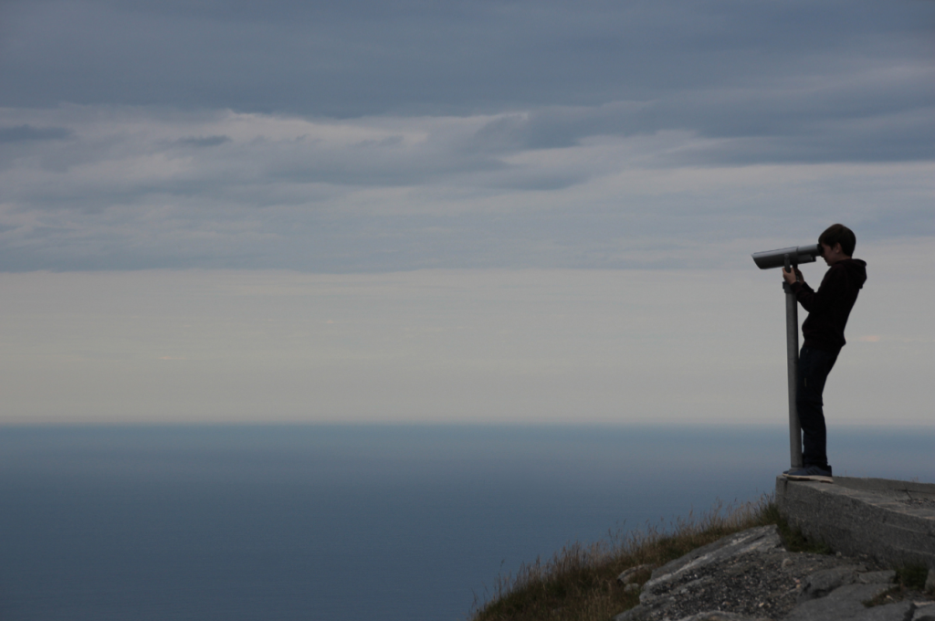 Mensch blickt mit Fernglas von einer Anhöhe auf das Meer hinaus.